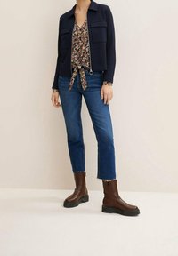 Giacca navy con zip e due tasche sul petto indossata sopra una blouse floreale con nodo davanti. Jeans blu a vita alta e stivali chunky marroni completano l'outfit.