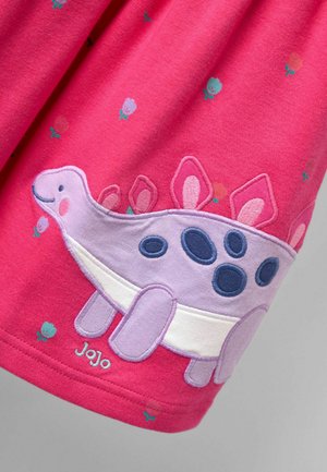 Rozā audums ar maziem ziedu rakstiem un smaidīga purpura dinozaura aplikācija ar ziliem punktiem un mīkstiem ērkšķiem. Zemāk izšūts vārds "JoJo".