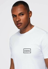 Ung mand med kort hår iført en hvid t-shirt med et sort "OMBRE" logo på brystet, der kigger direkte ind i kameraet.