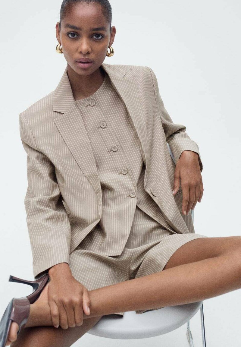 Completo beige a righe con giacca su misura e pantaloni corti abbinati. Caratterizzato da tre bottoni e una texture morbida. Abbinato a tacchi scuri.