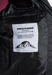 Crna unutarnja obloga jakne, sa bijelom etiketom na kojoj je logo "PEGADOR", grafika planine i upute za njegu otisnute crnim tekstom.