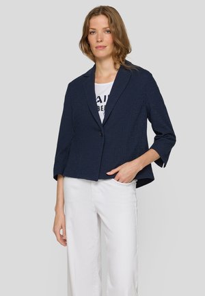Frau trägt einen marineblauen Blazer über einem weißen Hemd mit Print und weißen, weit geschnittenen Hosen, steht mit einer Hand in der Tasche vor einem grauen Hintergrund.