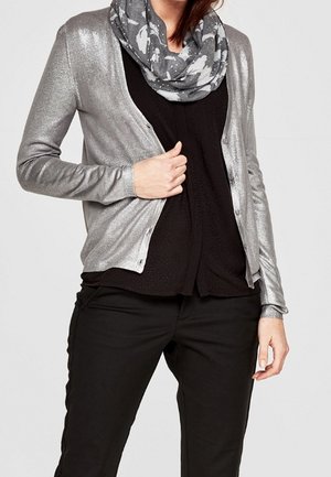 Vrouw draagt een metalen zilverkleurig vest over een zwart shirt, gecombineerd met zwarte broek en een grijs patroon sjaal om de nek.