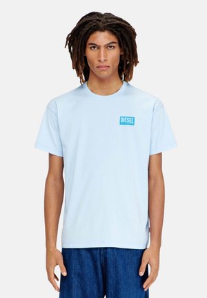 Giovane con dreadlocks di media lunghezza che indossa una maglietta Diesel azzurro chiaro e jeans blu scuro, in piedi davanti a uno sfondo uniforme.