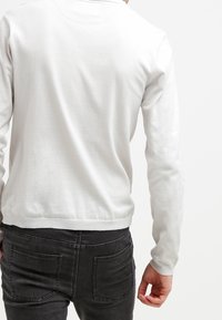 Biały sweter z długim rękawem, wykonany z miękkiego materiału, z okrągłym dekoltem i ściągaczami na rękawach. Widok z tyłu ukazuje dopasowany krój z gładką fakturą.