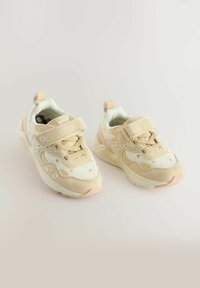 Beige sportsko med mesh- og ruskindsmateriale, der har et blomstermotiv, dobbelt velcro-lukning og en polstret sål.