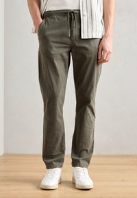 Pantaloni din bumbac ușor, de culoare verde măsliniu, cu talie elastică și șnur. Au buzunare laterale și picioare îngustate. Purtați cu adidași albi.