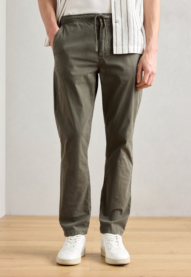Pantaloni din bumbac ușor, de culoare verde măsliniu, cu talie elastică și șnur. Au buzunare laterale și picioare îngustate. Purtați cu adidași albi.