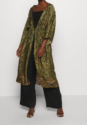 Frau trägt grünen Paisley-Kimono mit gelbem Muster am Saum über schwarzem Tanktop und weiten Hosen, steht mit einer Hand nach unten.