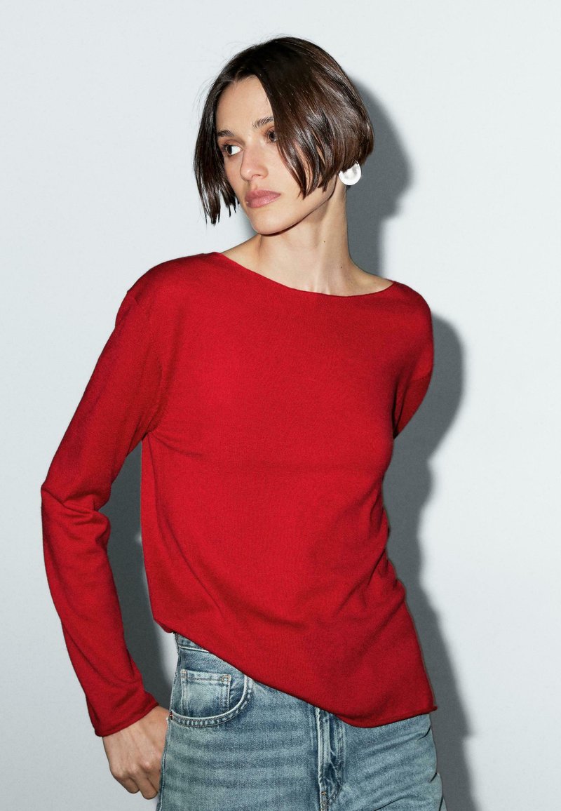 Model wearing a red long-sleeve top and denim jeans, posing against a light gray background, with a thoughtful expression and short hair.