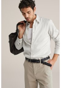 Mann in weißem Nadelstreifenhemd und beiger Hose, der eine schwarze Ledertasche über der Schulter trägt, schaut nach unten und hat eine Hand in der Tasche.