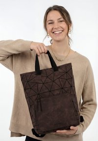 Usměvavá žena v béžovém svetru drží hnědou tašku s geometrickým vzorem, černými úchyty a přední kapsou na zip.