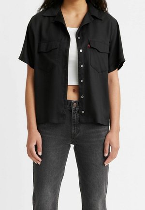 Kobieta ubrana w czarną, krótkim rękawem, rozpiętą koszulę na guziki, na białym, krótkim topie oraz w wysokowyszarzane, czarne dżinsy z wysokim stanem.