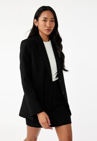 Blazer negro entallado con un ajuste estructurado, cierre de un solo botón y dos bolsillos frontales, usado sobre una blusa clara y una falda negra texturada.