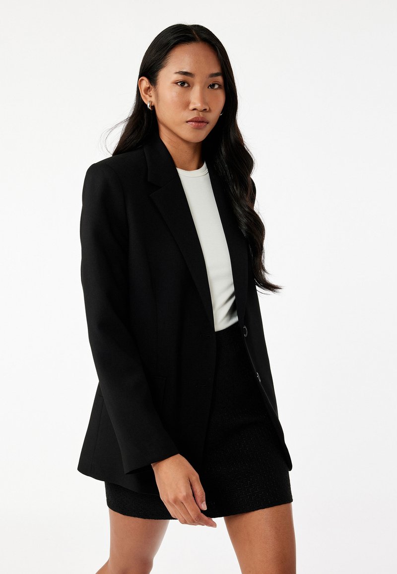 Schwarzer taillierter Blazer mit strukturiertem Schnitt, einfacher Knopfverschluss und zwei Fronttaschen, getragen über einem hellen Oberteil und einem strukturierten schwarzen Rock.