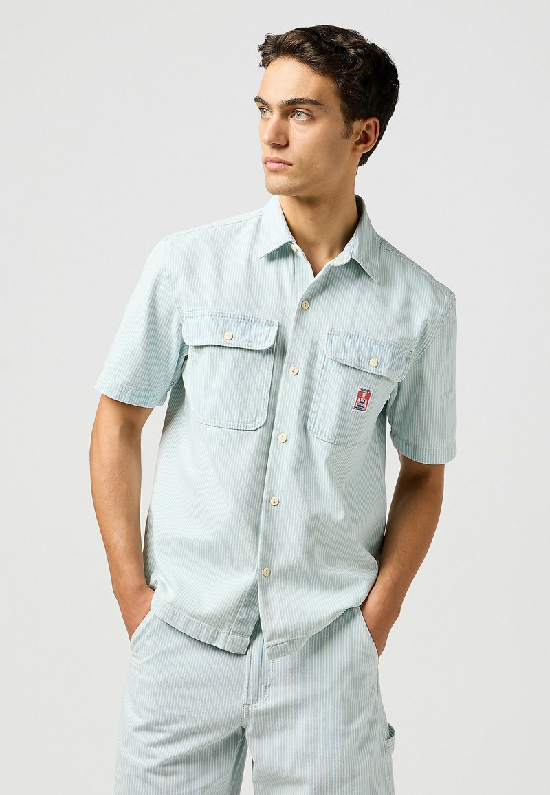 Lichtblauw kortouwen overhemd met verticale witte strepen, twee borstzakken en knoopsluiting. Glad materiaal met een comfortabele pasvorm.