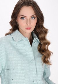 Sacou din tweed de culoare albastră deschisă, cu un model texturat, design cu guler, nasturi în față și un buzunar pe piept. Include detalii elegante de cusătură.