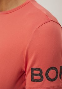 T-shirt de sport rouge avec une texture lisse et des manches courtes. "BORN" imprimé en noir avec un motif en pointillés sur la manche droite.