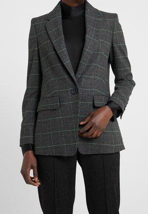 Blazer à carreaux gris foncé avec des lignes vertes subtiles porté sur un haut noir et associé à un pantalon noir, main ajustant le blazer.