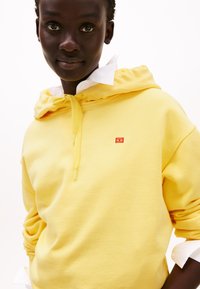 Felpa gialla con zip frontale, caratterizzata da un piccolo logo rosso. Il tessuto sembra morbido, sovrapposto a una camicia bianca con colletto.