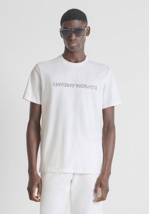 Hombre con gafas de sol negras, camiseta blanca de Antony Morato y pantalones blancos frente a un fondo claro y liso.