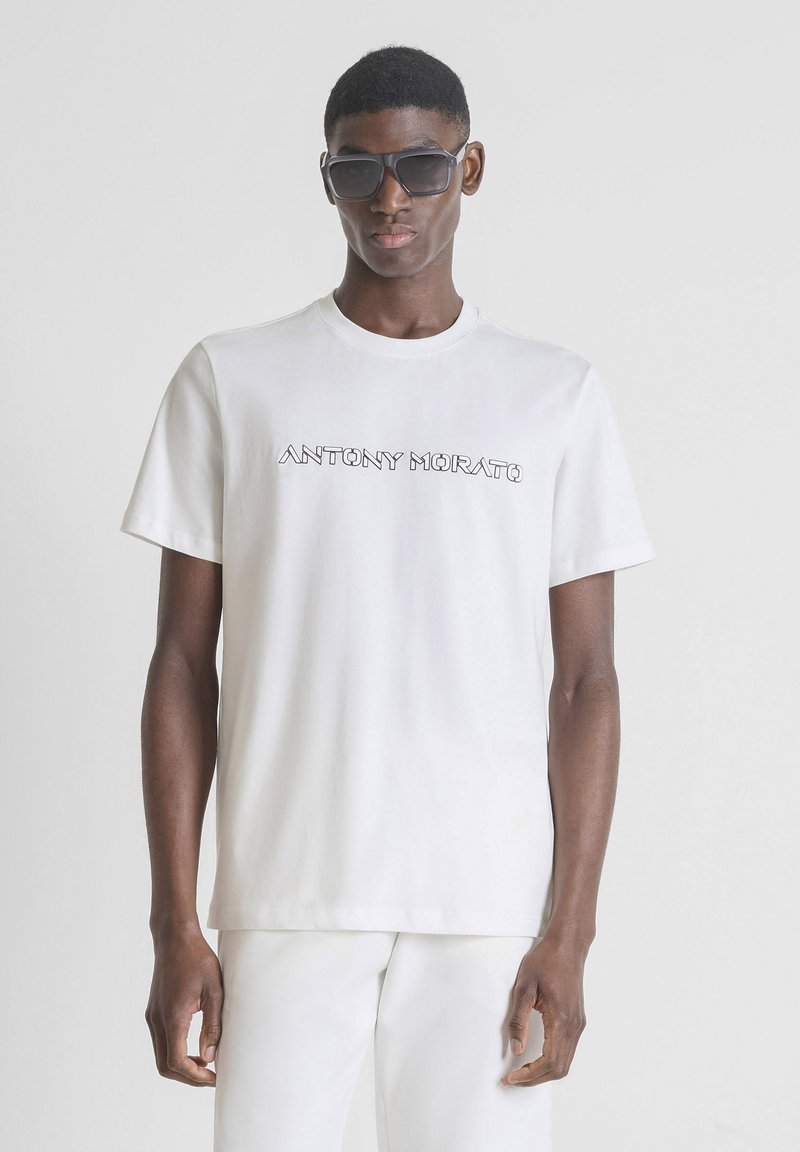 Hombre con gafas de sol negras, camiseta blanca de Antony Morato y pantalones blancos frente a un fondo claro y liso.