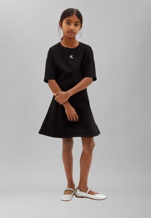 Jeune fille debout, les bras croisés, vêtue d'une robe noire à manches courtes et de chaussures plates blanches sur un fond gris uni.