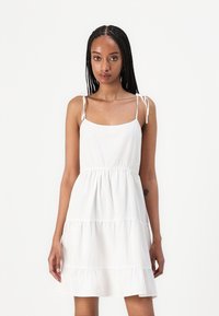 White tiered dress made of lightweight fabric, featuring adjustable spaghetti straps and a gathered waist for a relaxed fit.