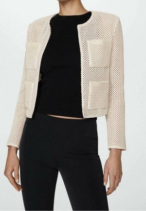 Veste beige courte en maille ouverte avec manches longues et deux poches plaquées devant, portée sur un top noir ajusté et un pantalon noir.