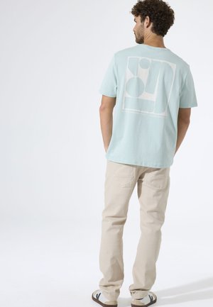 Mintgroen katoenen T-shirt met een grote abstracte witte afbeelding op de achterkant. Gecombineerd met lichtbeige broek en casual schoenen.