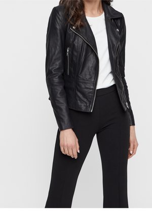 Femme portant une veste en cuir noire zippée sur une chemise blanche et un pantalon noir, debout les bras détendus devant un fond uni.