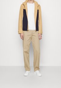 Lys gul og marineblå jakke med hætte, parret med beige bukser og hvide sneakers. Stoffet ser glat ud med rene linjer.