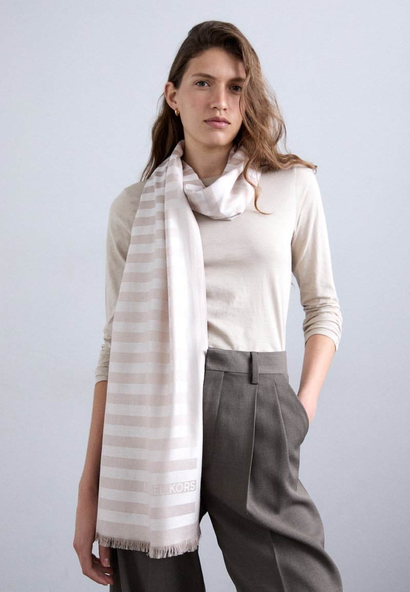Écharpe rayée rose clair en tissu doux, dotée d'un ourlet à franges et d'un accent logo subtil, enroulée autour d'un haut neutre.