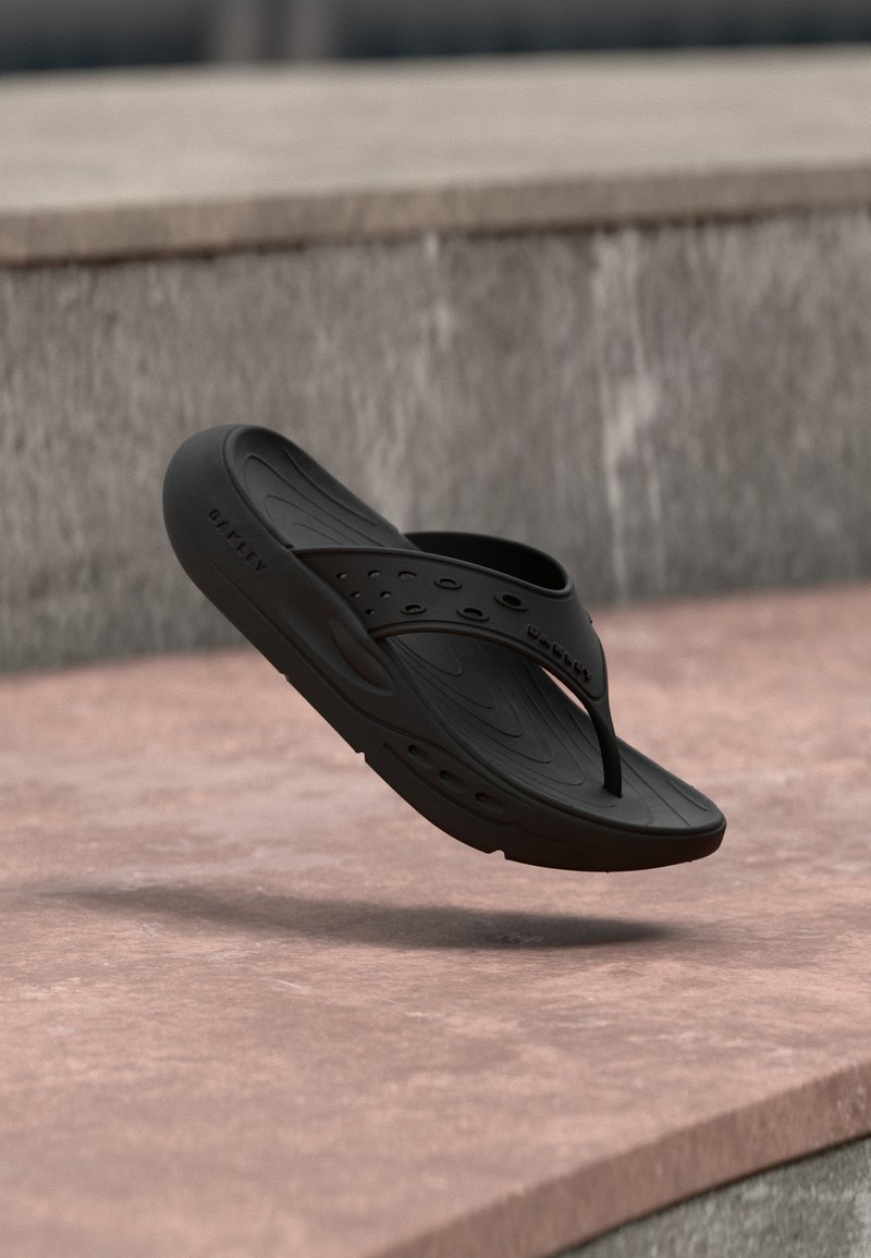 Μαύρο λαστιχένιο σαγιονάρι με τρύπες και υφή στην σόλα, αιωρείται πάνω από ροζ επιφάνεια από σκυρόδεμα και γκρι πέτρινο φόντο.