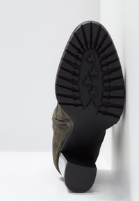 Semelle noire et talon de bottine en daim vert avec fermeture éclair sur le côté, présentée debout contre un fond blanc en coin.