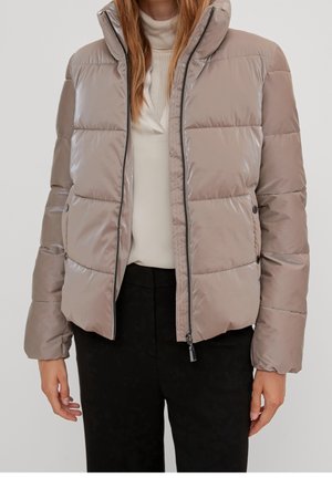 Woman wearing a light beige puffer jacket over a white turtleneck and black pants, standing against plain background.