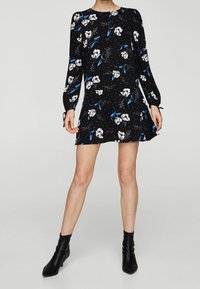Robe noire à manches longues, ornée d'un motif floral blanc et bleu, jupe évasée et col rond. Portée avec des bottines noires.
