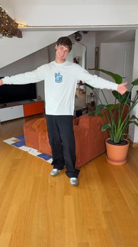 Bijela košulja dugih rukava s plavom grafikom, crne široke hlače i srebrne cipele. Unutarnji prostor s biljkom i smeđim kaučem s prugama.