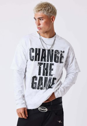 Jeune homme aux cheveux décolorés portant une chemise blanche à manches longues avec le texte « CHANGE THE GAME » et des chaînes argentées superposées, regardant sur le côté.