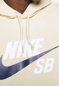 Närbild av en beige hoodie med vita snoddar och en stor marinblå Nike swoosh och "NIKE SB"-text på bröstet.