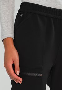 Svarta träningsbyxor med ribbad midja, dragkedjeficka på sidan och slät textur. Handen vilar på tyget.