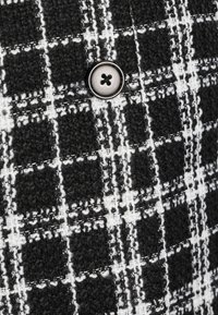 Musta ja valkoinen ruutukuvioinen kangas, jossa on karhea pinta, ja etualalla metallinen nappi, jossa on neljä reikää.