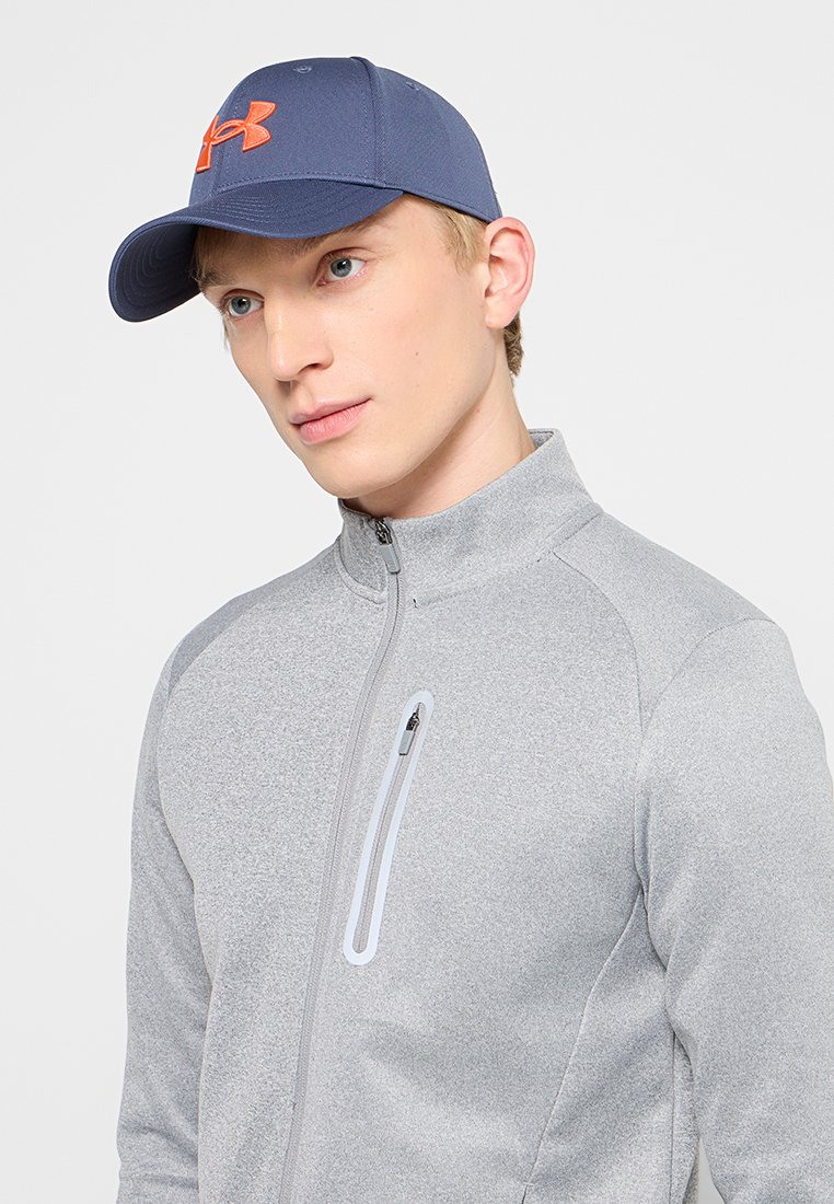 Blaues Cap mit einem orangefarbenen Logo, kombiniert mit einer hellgrauen, taillierten Zip-Jacke mit einem Reißverschlussfach und strukturiertem Stoff.