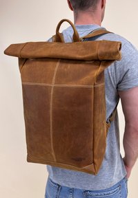 Ruda odinė ritininė kuprinė su balta siūle, stačiakampio formos ir viršuje turinti rankeną. Apatinėje pusėje yra logotipas.