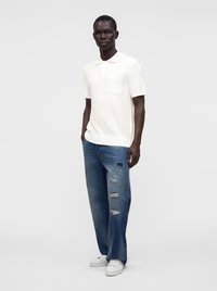 Homme portant un polo blanc texturé à manches courtes, un jean large bleu déchiré et des baskets blanches, debout devant un fond uni.