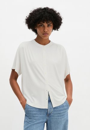 Woman with curly hair wearing a short-sleeve white button-up blouse and light blue jeans, hands in pockets, neutral expression.