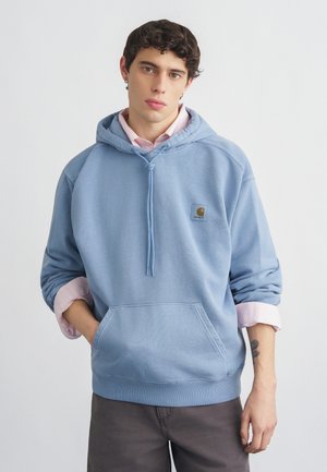 Fiatal férfi rövid, göndör hajjal, világoskék kapucnis pulóvert visel felhajtott rózsaszín ing fölött, egyik kezével a zsebében áll, fehér háttér előtt.