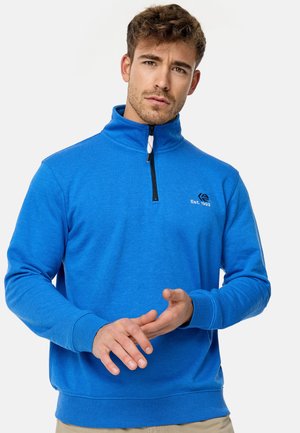 Mann mit lockigem braunem Haar, der einen hellblauen Sweatshirt mit Viertelreißverschluss trägt, berührt seine Hände und schaut leicht ernst vor einem einfarbigen Hintergrund.