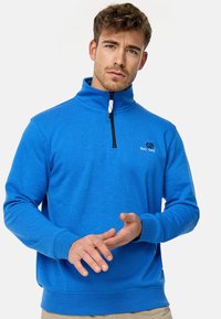 Lys blå fleece-pullover med høj krave, halvdækkende lynlås og sort lynlås. Har et lille logo på brystet og ribbede manchetter.