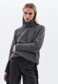 Maglione grigio a collo alto con colletto e polsini a coste, realizzato in un materiale morbido e testurizzato. Abbinato a pantaloni neri lucidi a vita alta con dettagli laterali.